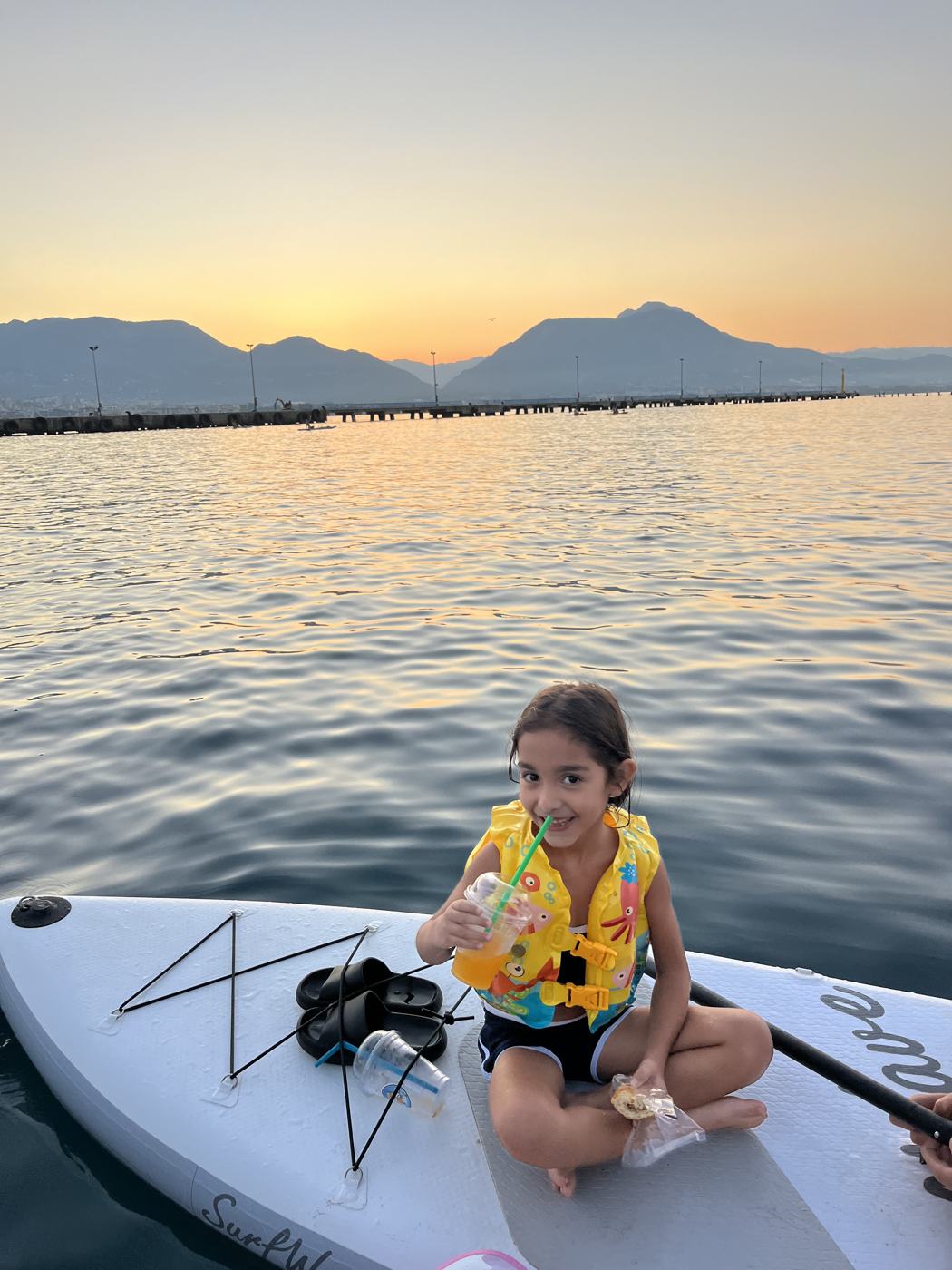Child in a life jacket sitting on a board at sunrise